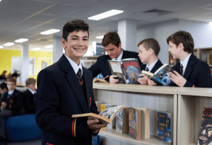 A photo of a secondary school student smiling with a book while his three friends in the background also read